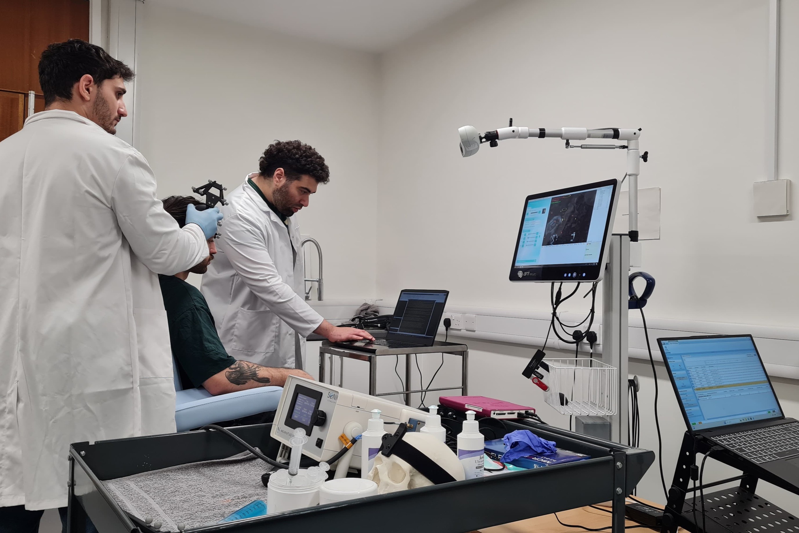 Two researchers in white lab coats, look at a computer screen with data whilst a patient receives a treatment