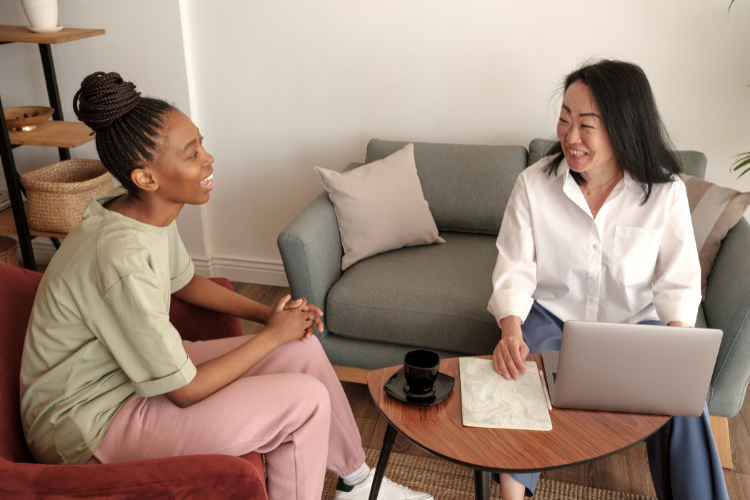 Two people are in conversation. The one on the left is wearing a grey tshirt and pink trousers, the one to the right is in a white shirt sat at a laptop