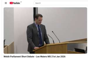 A screenshot of an online video featuring a man standing at a microphone addressing a room full of people